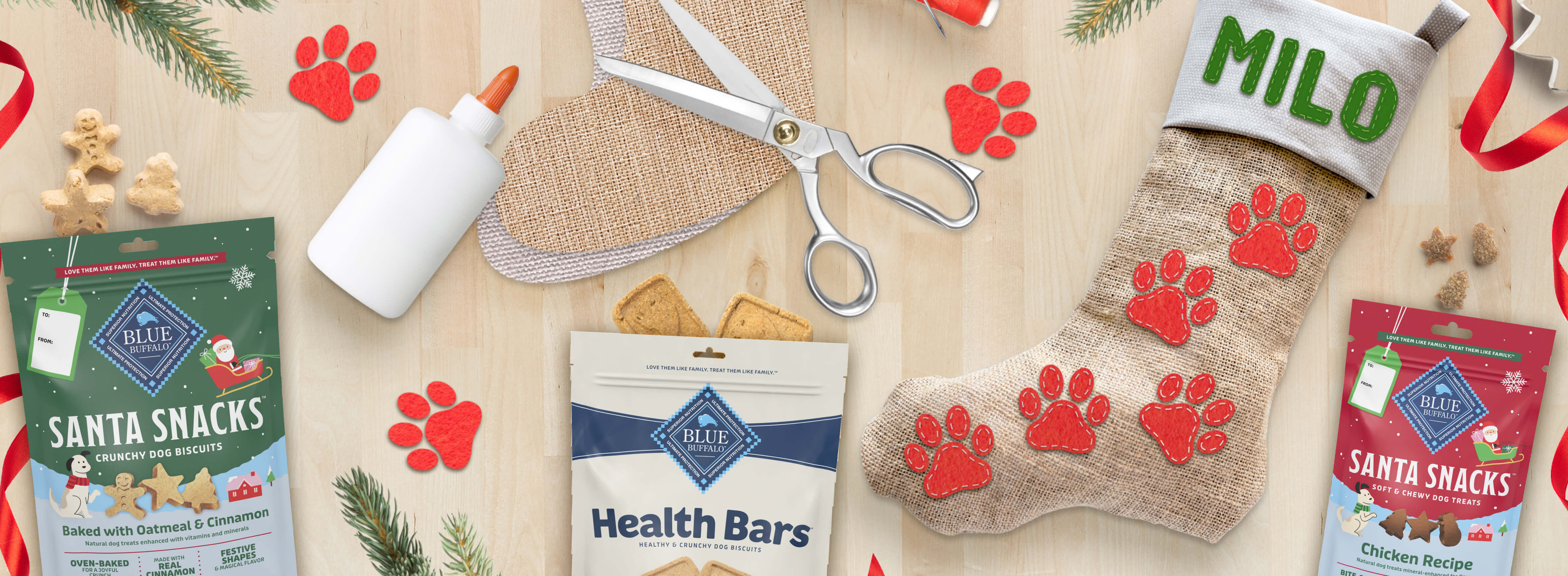 Dog treat packages, scissors, glue, stocking labeled 'Milo', and holiday decorations on table.