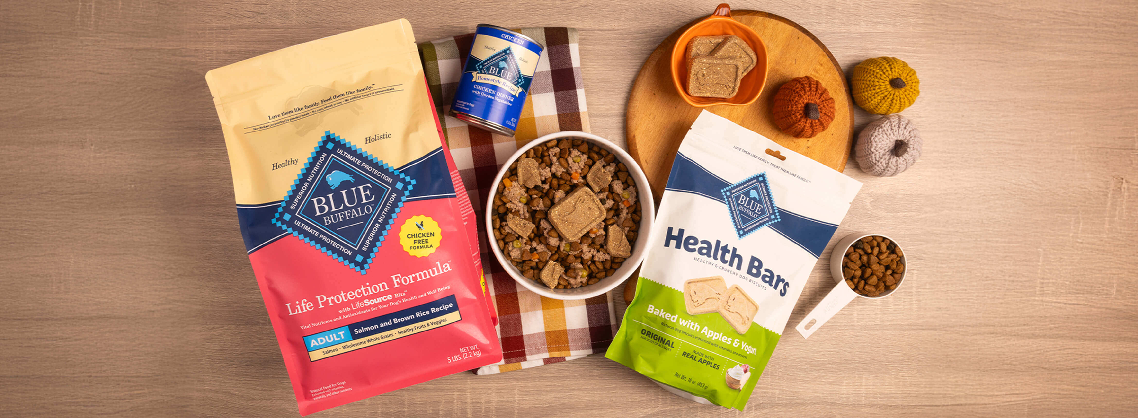 A dog-friendly meal sits in a bowl on a hand towel, surrounded by packages of BLUE Life Protection Formula, BLUE Homestyle Recipe, and BLUE Health Bars, along with autumnal decor.