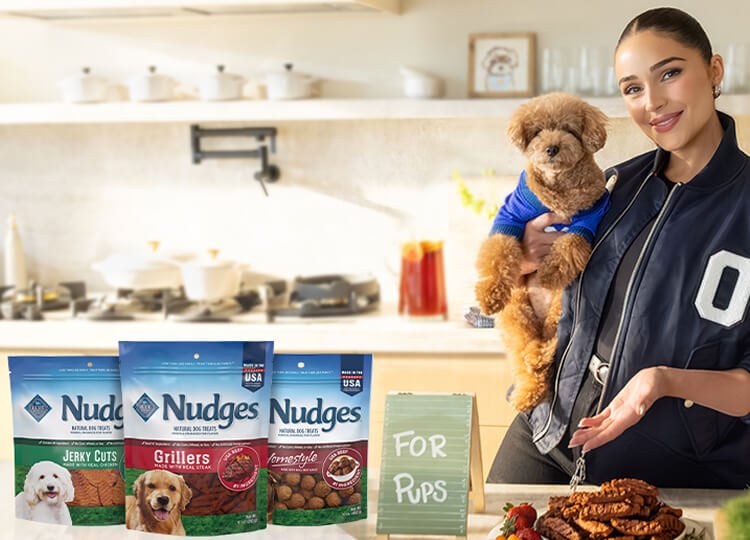 A woman holds a small dog in a kitchen with Nudges dog treats and a sign that reads "For Pups" on the counter.