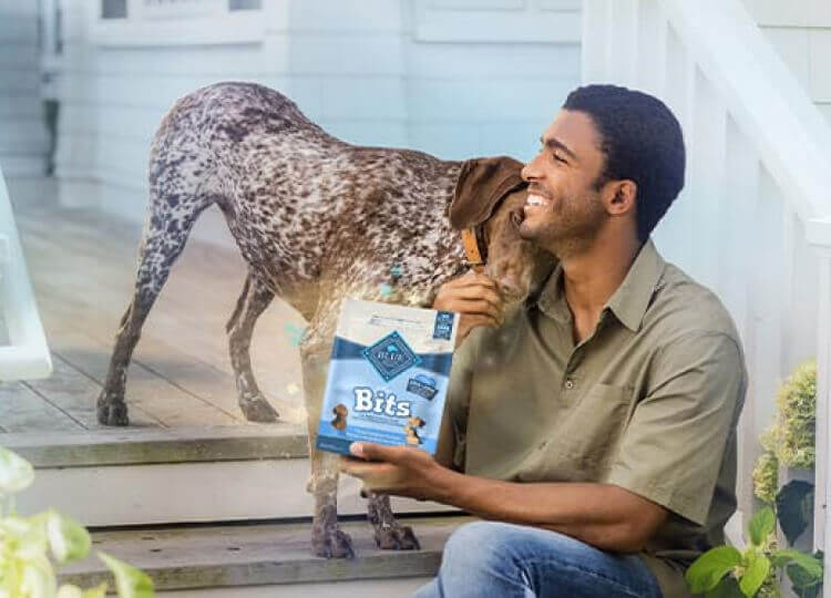 A man is feeding his dog a treat. A bag of BLUE Bits dog treats is in his other hand. They’re sitting on the steps of a porch.